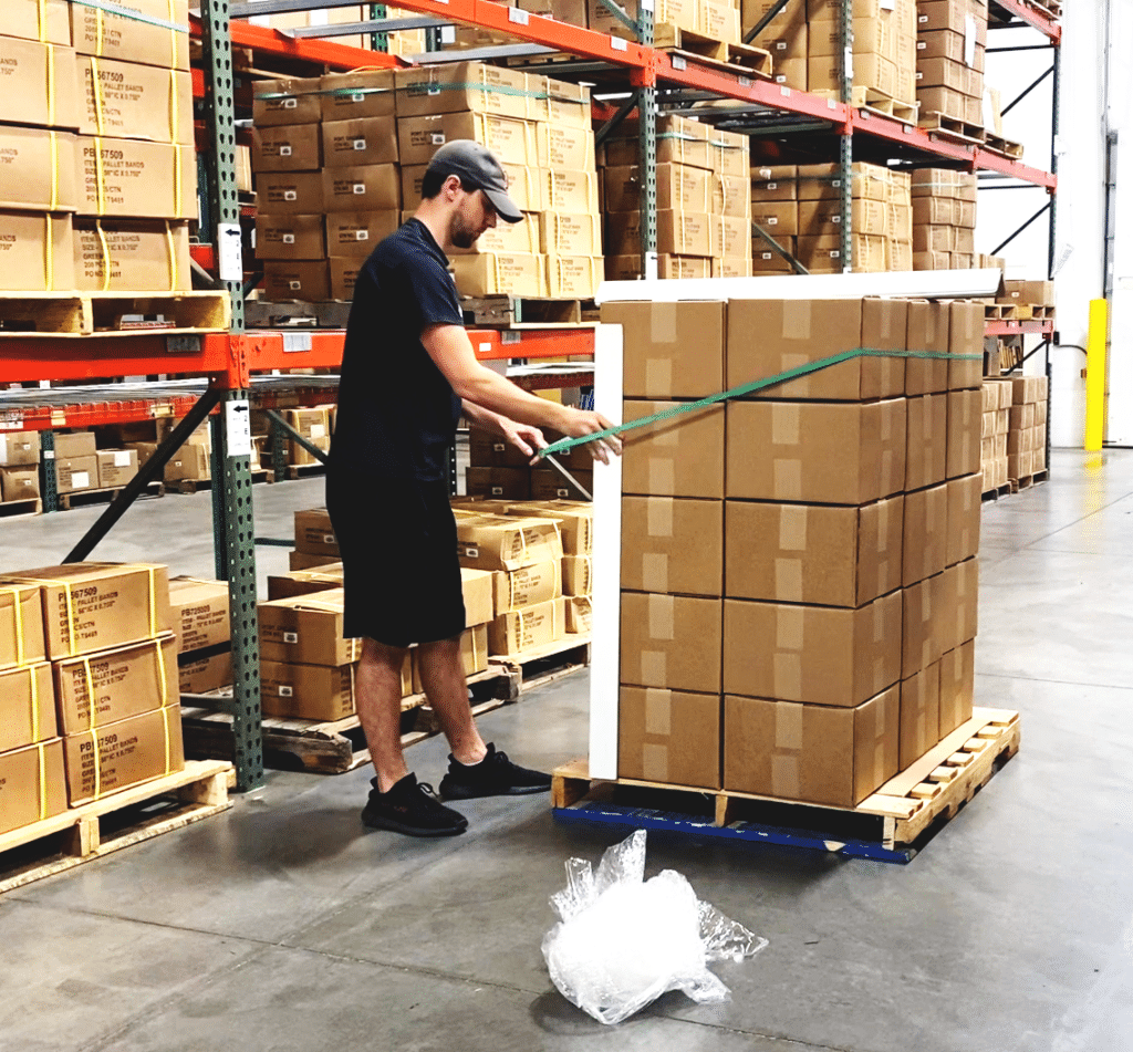A young man applying a large rubber band on a pallet of cartons in a warehouse.